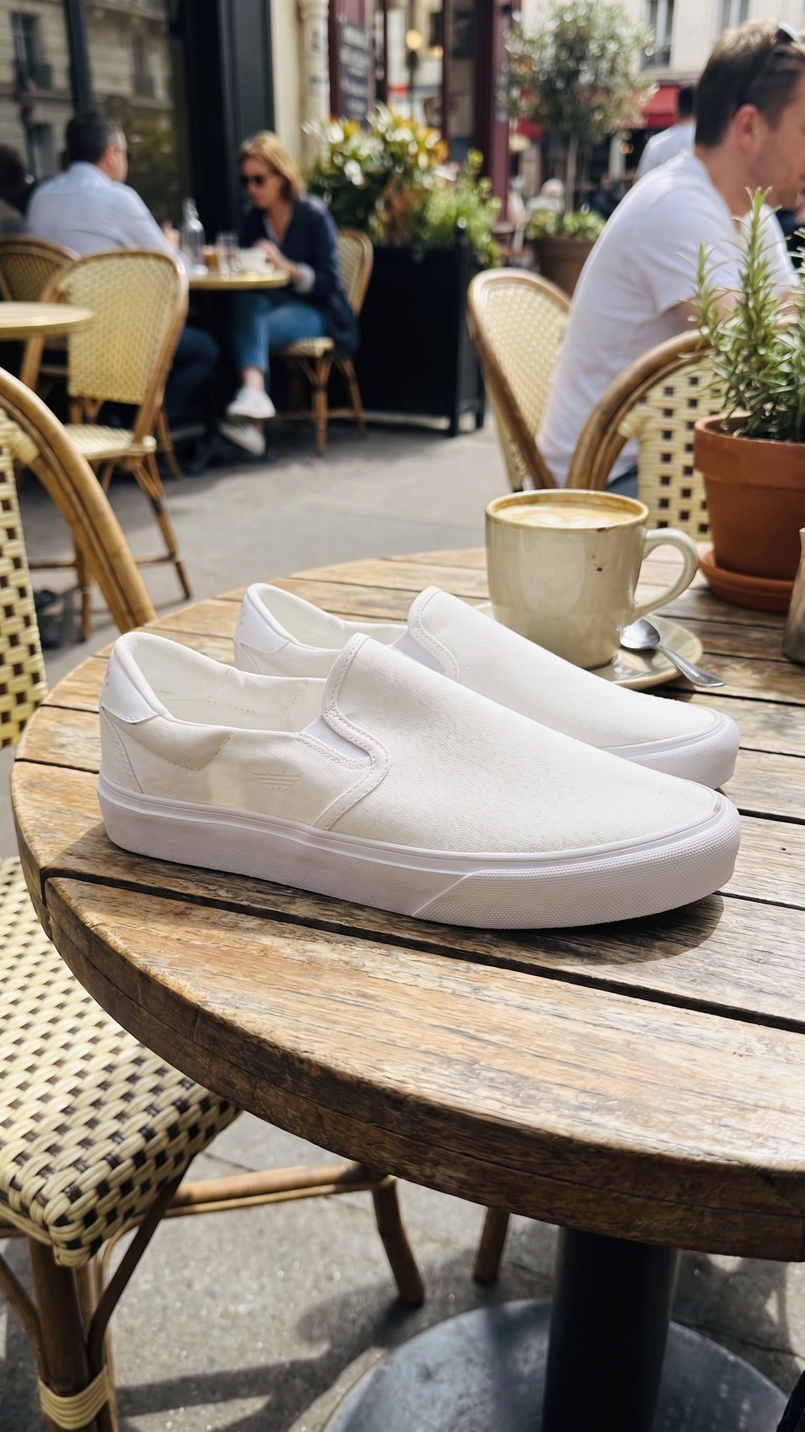 Two white slip-on sneakers resting on a wooden outdoor cafe table.