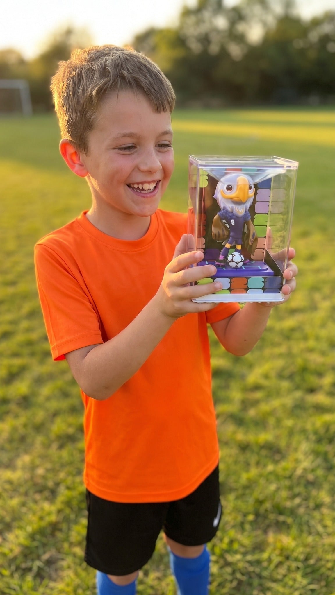 A boy in a football kit holding the cased Clutch the Bald Eagle figurine on a sunlit pitch