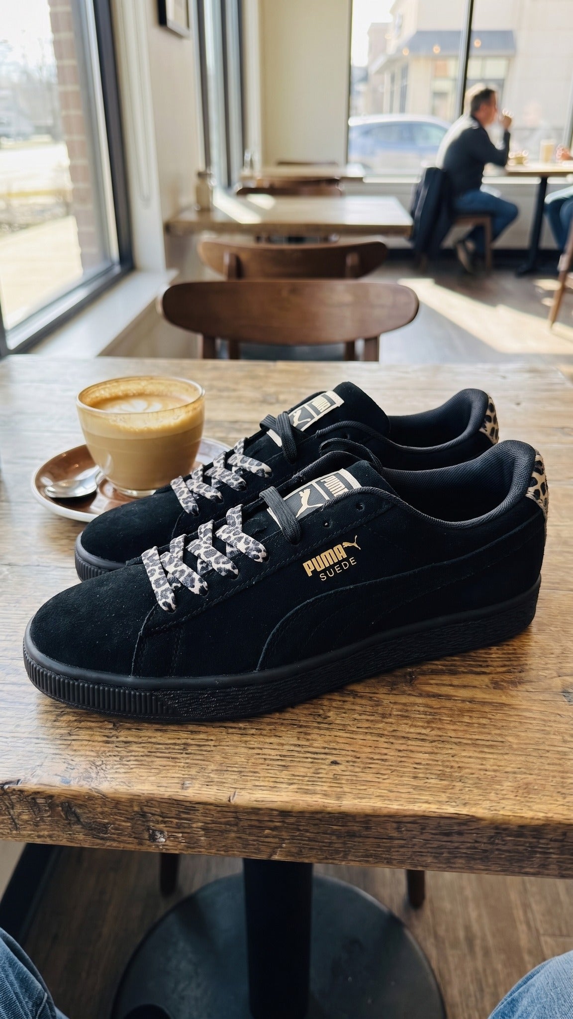 Both shoes sitting on a wooden table in a cafe next to a cup of coffee.